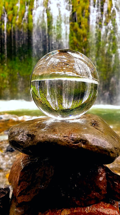 Glaskugel auf Steinen spiegelt Wasserfall und herbstliches Laub wider.