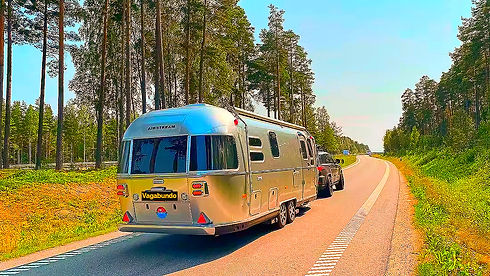 Silberner Wohnwagen auf Landstraße, umgeben von Wald, sonniger Tag.