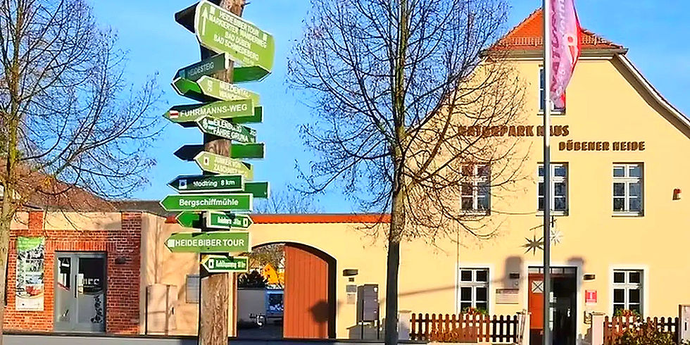 Wegweiser mit vielen Schildern, Naturpark Haus Dübener Heide, gelbes Gebäude im Hintergrund.