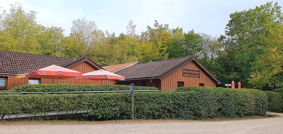 Holzgebäude des Besucherzentrums Teufelsschlucht hinter Hecken, mit roten Sonnenschirmen und grünem Wald im Hintergrund.