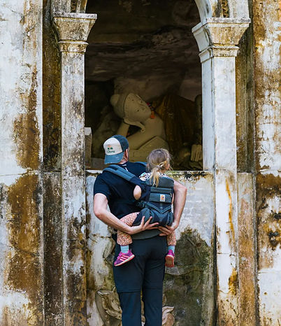 Elternteil trägt Kind in Rückentrage vor Ruine.
