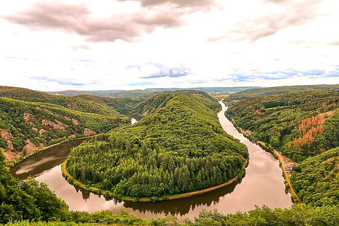 Breiter Flussmäander inmitten grüner bewaldeter Hügel unter bewölktem Himmel.