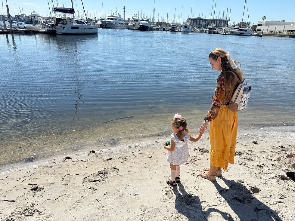 Woman and child holding hands on a sandy shore by a marina. Boats are moored in the background. The woman wears a yellow skirt. Sunny day.