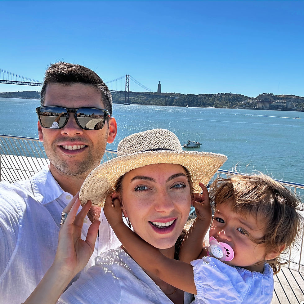 Family selfie by a sunny waterfront with a bridge in the background. Woman in straw hat smiles, child with pacifier embraces her.
