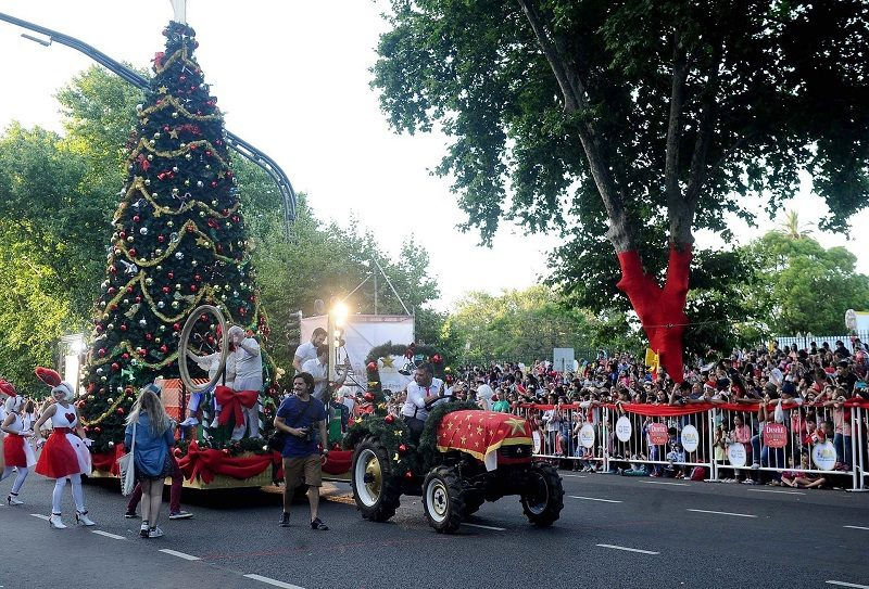 Christmas parade with a decorated tree float and people in festive costumes. A crowd watches along the street. Bright, cheerful atmosphere.