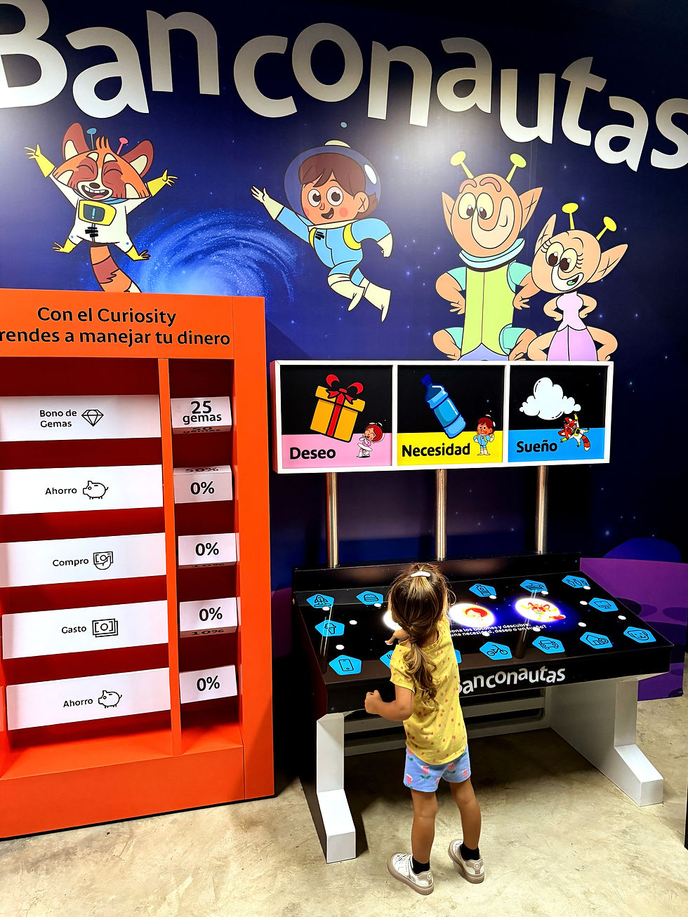 A toddler girl interacts with the Banconautas financial literacy exhibit at Explora Science Center in Panama City, Panama, featuring colorful cartoon characters and a touchscreen table.