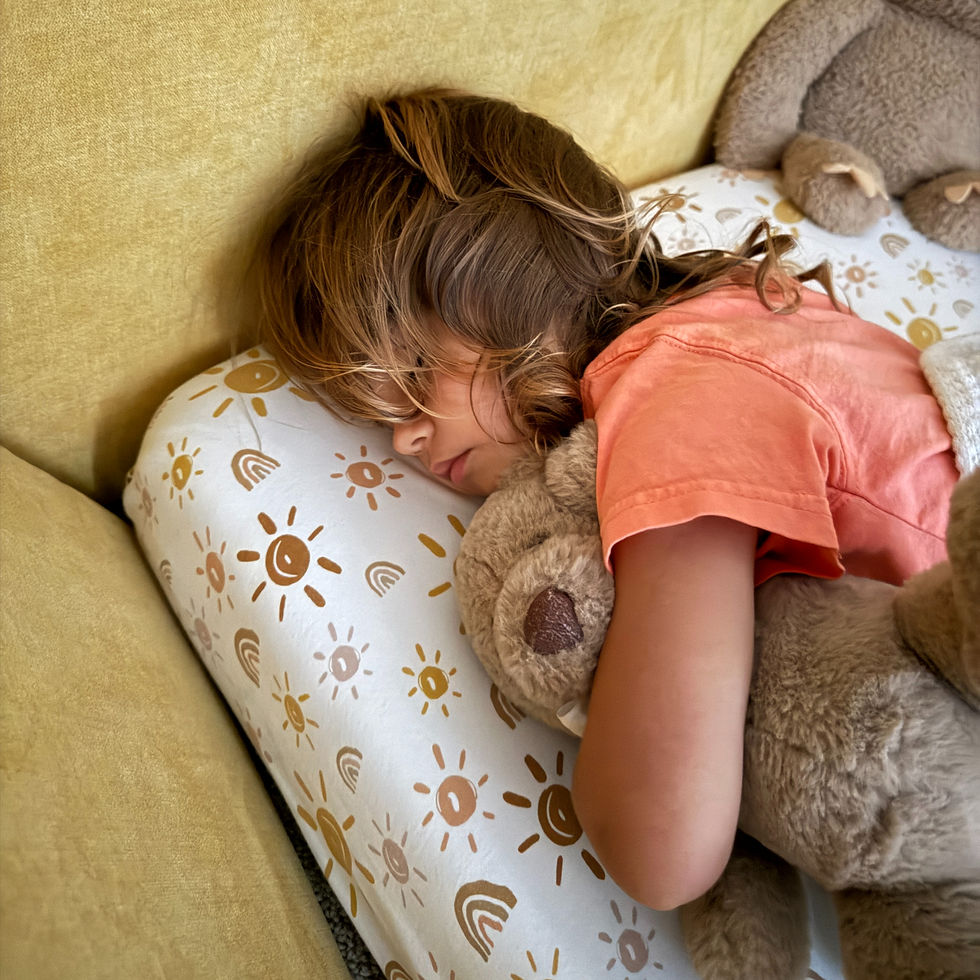 Child sleeping on a sofa, hugging a teddy bear. Wears an orange shirt. Pillow has a sun pattern. Calm and cozy atmosphere.