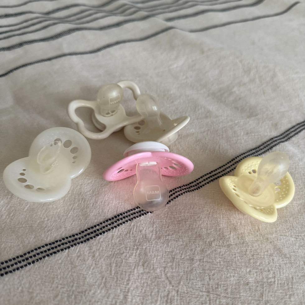 Four pacifiers on a striped fabric, with one pink and the others white and yellow. The word "MAM" is visible on the pink one.