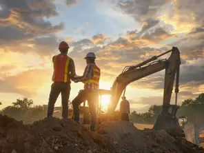 Two construction workers at a site, backlit by a sunset with an excavator behind them.