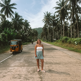 Meisje met groene bloemetjesrok en witte top staat midden op een weg met daarlangs veel palmbomen en links van haar een gele tuktuk.