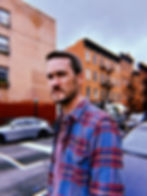 A photo of Zach Powers, a skinny, middle-aged white man with brown hair and beard. He’s standing on a sidewalk in Brooklyn with brownstones in the background. He’s wearing an
unbuttoned blue and red flannel shirt over a red Henley.