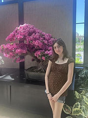A young white woman with medium length dark brown hair smiles at the camera. She wears a brown babydoll top with flowers and stands in front of a pink bonsai tree.