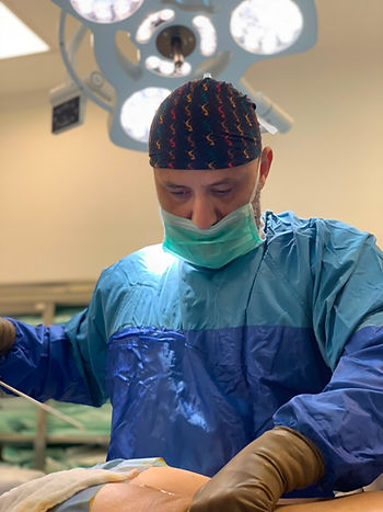 Surgeon in blue gown and mask works over a patient in an operating room under bright surgical lights.