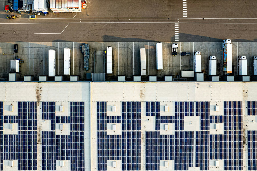 Aerial view of a large logistics warehouse with a rooftop solar array, trucks at the loading docks and a parking lot