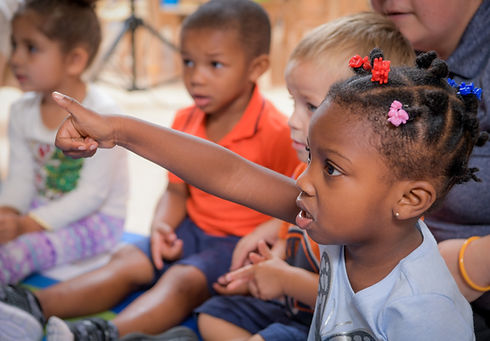 A group of preschool aged children sit together. One holds her hand up, thumb raised