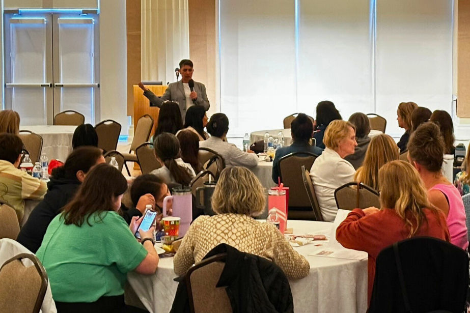 A speaker presents to an attentive audience in a conference room with round tables. Attendees sit, some taking notes, in a well-lit setting.