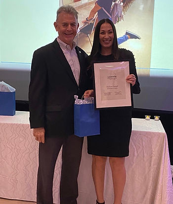 A man and woman stand in front of a table. The woman holds a gift bag and certificate.