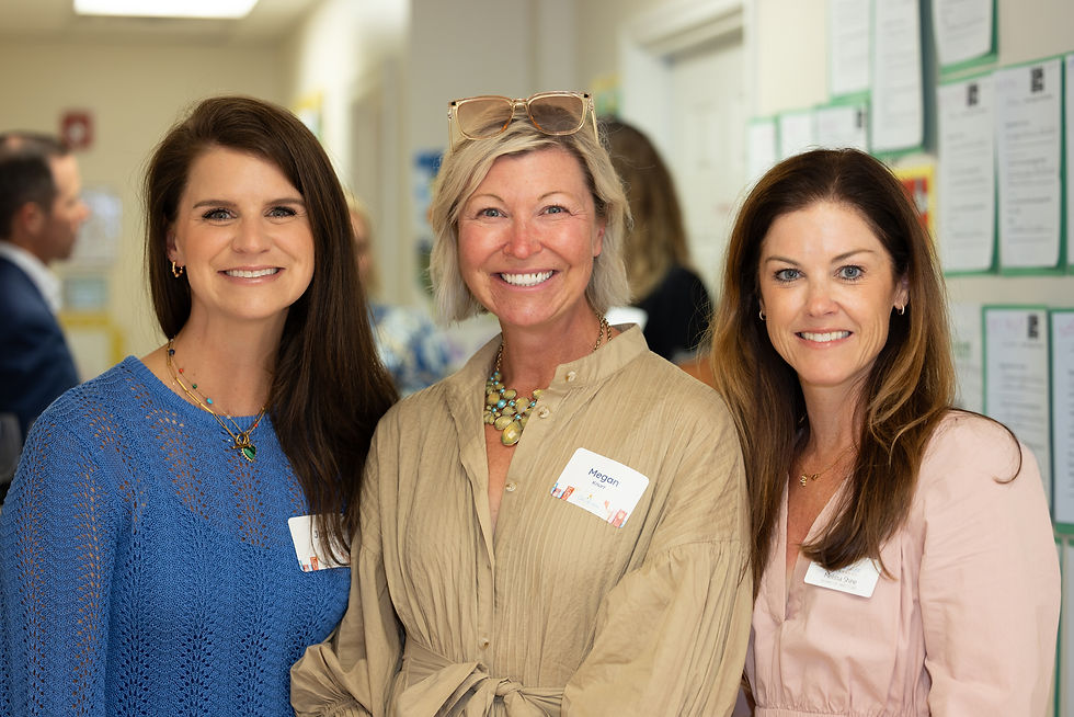Three women smiling in a hallway, one wearing a blue top, another beige, and the third pink. Name tags visible; bright, cheerful setting.