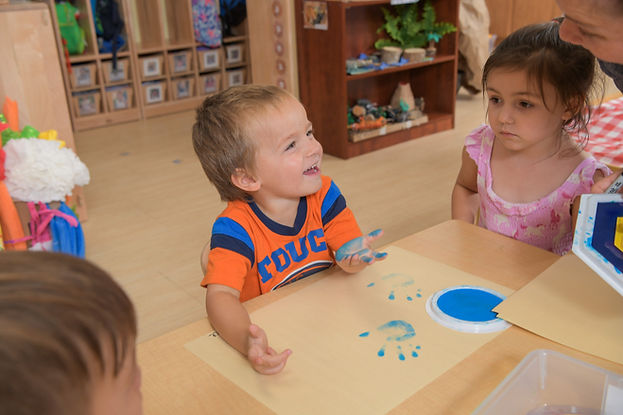 Two children sit at a table, fingerpainting with blue paint