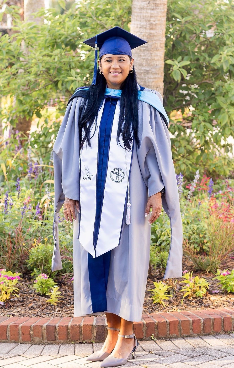 Smiling graduate in blue cap and gray gown with UNF stole stands outdoors in a garden.