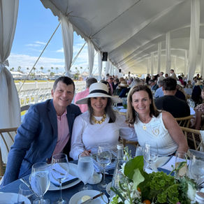 Three people smiling at a table under a white tent, outdoors. There are glasses, plates, and green plants on the table. Blue sky outside.