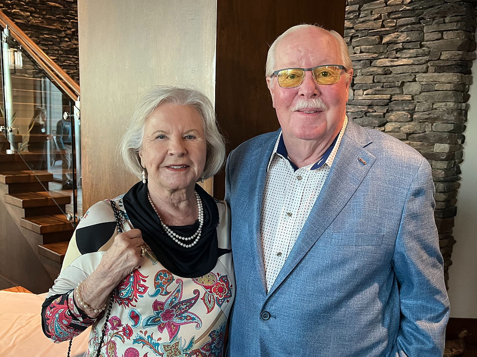 A couple smiles in a warm setting with stone walls and wood stairs. The woman wears a colorful top, the man a blue jacket and yellow glasses.