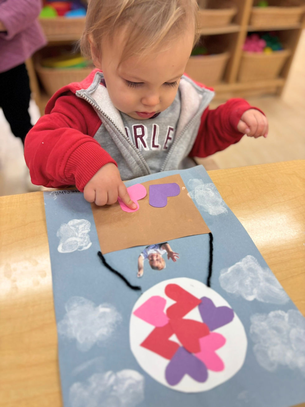 A toddler in a red jacket places heart shapes on a craft with a photo of themselves. The background is a classroom with shelves.