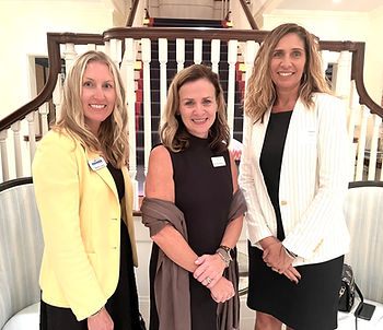 Three women smiling, wearing name tags, stand in front of a staircase. One in yellow, one in black, and one in a white jacket. Bright setting.