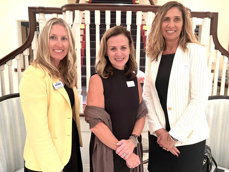 Three women smiling, wearing name tags, stand in front of a staircase. One in yellow, one in black, and one in a white jacket. Bright setting.