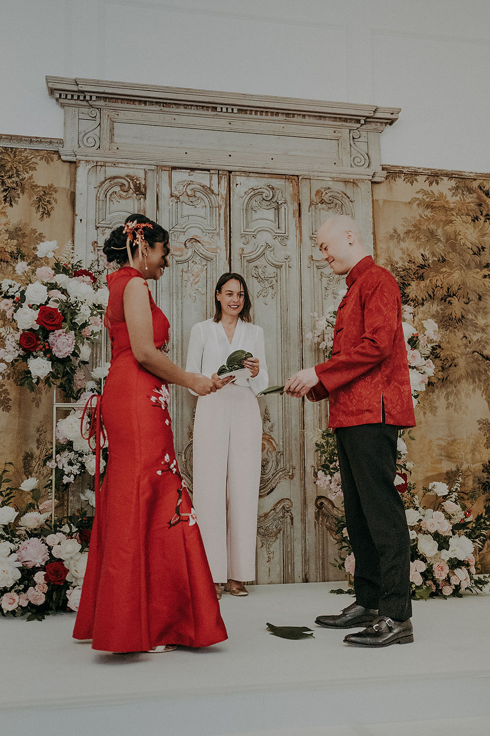 Wedding ceremony showing a bride in a red dress, a celebrant in a light pink jumpsuit and a groom in. a red jacket and black trousers on a stage with a panelled backdrop decorated with white, red and pink flowers. The couple and celebrant are holding big green leaves.