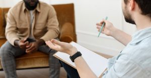 A man with a clipboard gestures while sitting across from a man. This could represent the support that somatic experiencing in Baltimore, MD can offer for overcoming trauma. Learn more about how a trauma-informed consultant in Baltimore, MD can offer support today.