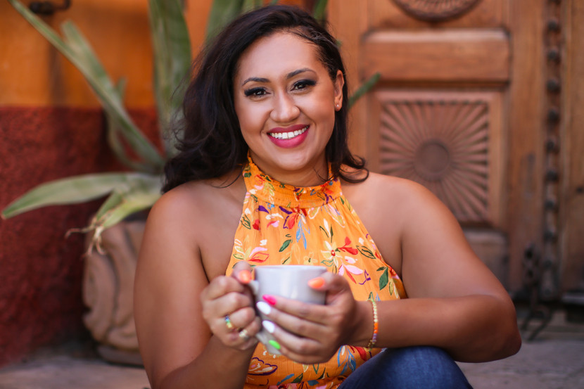 An astrologer sits on the steps in front of her door with a cup of coffee. She is smiling into the camera.
