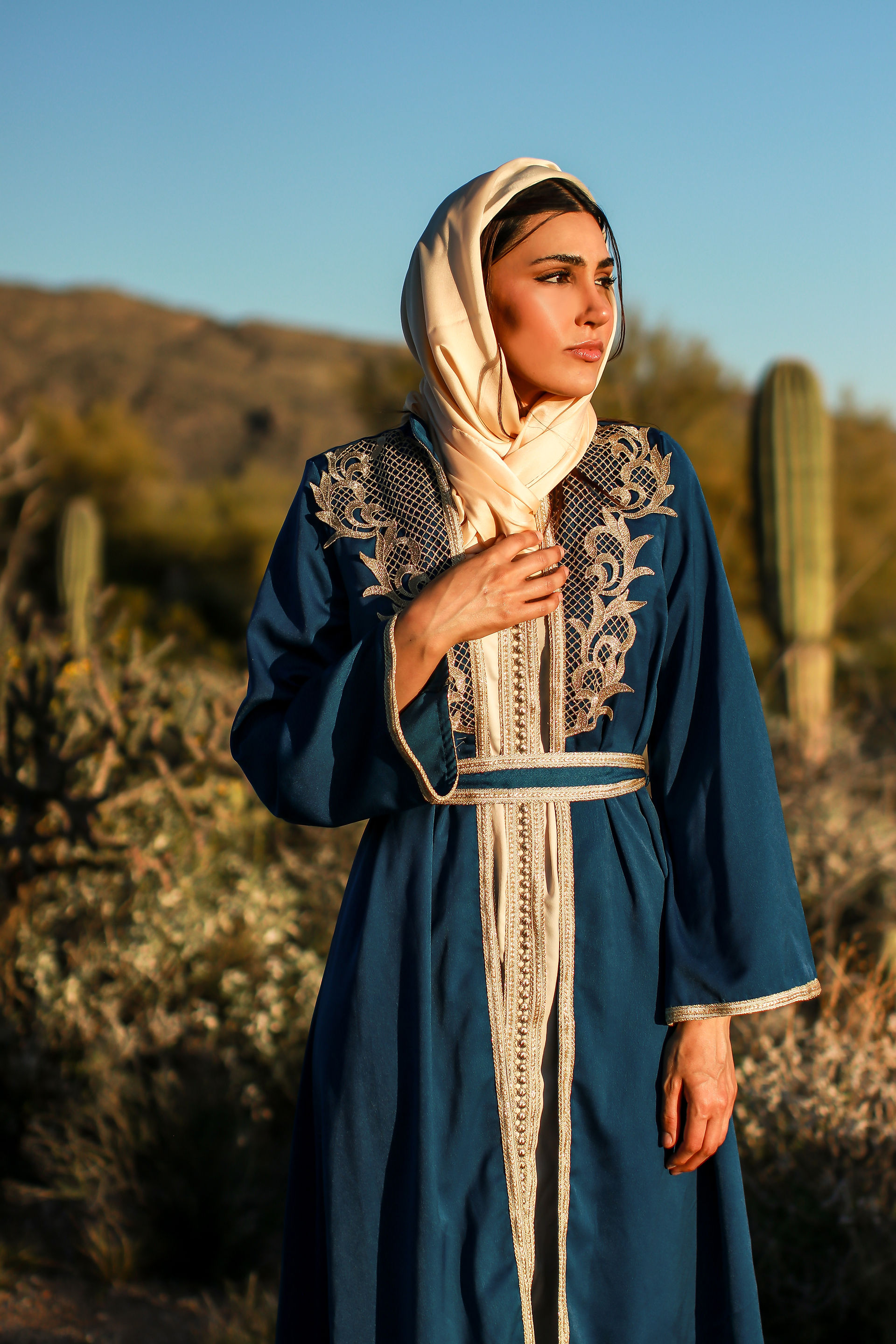 A woman walking in the Tucson desert turns to stare off toward the setting sun. She holds her abaya in place in the wind. 