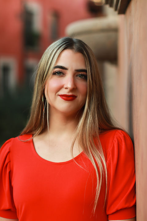 A young women with long blonde hair wearing a red dress and red lipstick faces the camera smiles for her closeup. She is looking straight into the camera as she leans against a wall. Grad Portrait of U of A Graduate by Tucson, AZ Grad Photographer Tina Lieberman . 