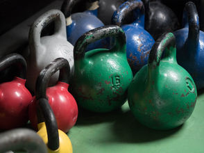 Colorful kettlebells in green, blue, red, yellow, and gray with visible wear, arranged on a green floor. Gym setting, no visible text.