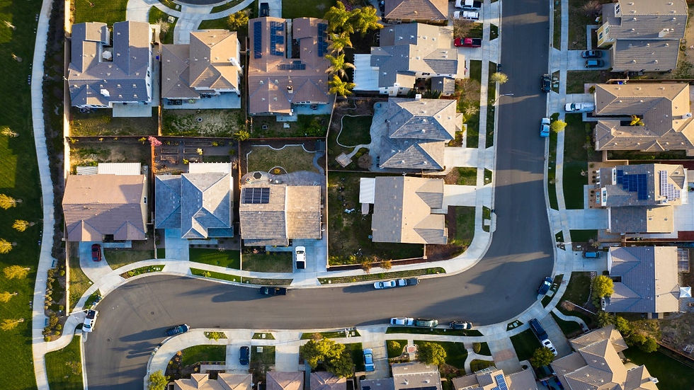 This is an aerial photograph of a suburban neighborhood at what appears to be either dawn or dusk, judging by the soft lighting. The layout of the community is clearly visible, with rows of closely situated single-family homes featuring variously pitched roofs. Some of the homes have solar panels installed. The neighborhood is well-planned with winding roads, neatly manicured lawns, and occasional backyard features like swimming pools and gardens. Vehicles are parked along the streets and in driveways, suggesting a peaceful residential area. The image captures the essence of suburban living, where each home sits snugly next to its neighbor, creating a cohesive community environment.