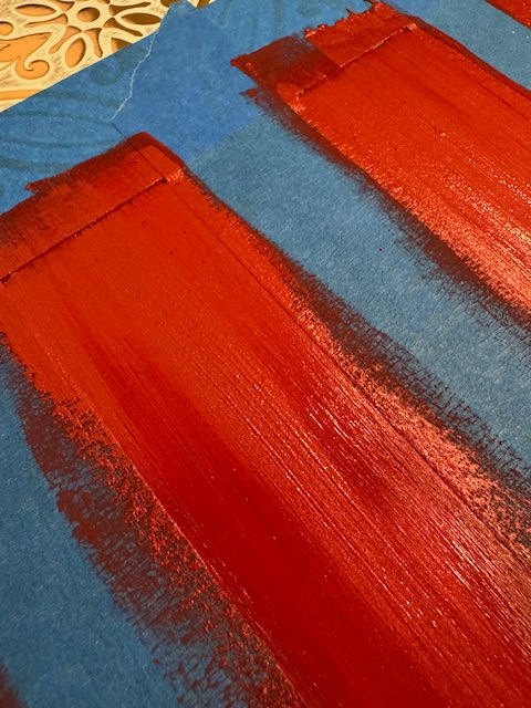 Bold red paint strokes on blue paper, with intricate tan patterns in the corner. The image has a dynamic and artistic feel.