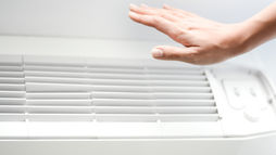 Hand near a white air conditioner, checking airflow in a bright, minimalistic room, highlighting the vents and controls of the best HVAC system.