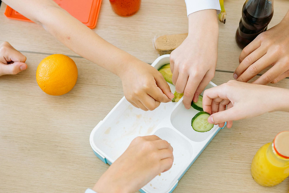 Hands reach for cucumber slices on a table with an orange, drinks, and a lunchbox, creating a communal, inviting atmosphere.