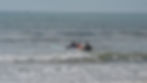 Três surfistas se preparam para entrar mais a fundo no mar. Dia de sol, dois surfistas com camisetas da pallet point surf e um menino com camisa e short laranja esta deitado na prancha sendo conduzido por outro surfista.