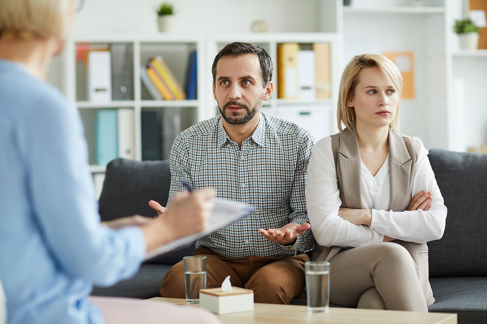 Serious couple in a counselling session on a sofa during family mediation in South Africa, with the man gesturing and the woman crossing her arms, shelves in background.