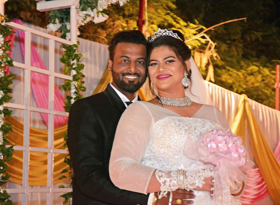 Amidst the "Sunset Serenade" inspired decor, an intimate moment unfolds with the bride CRYSTAL in her white gown, wearing a dazzling diamond necklace and holding a delicate bouquet, while the groom JOY, handsomely attired in a black suit, shares a loving pose at the entrance of thier wedding reception venue. Location IC colony , MUMBAI