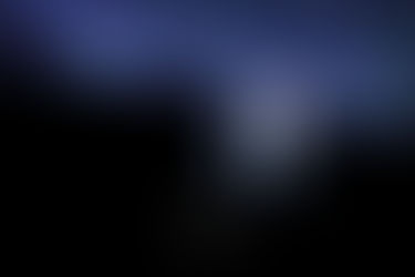 Shadows of a mountain range can be seen under a brilliant starry night sky. A woman stands on a rock and is illuminated from behind with a circle of light, her body and face in shadows, her arms poised like she is in the middle of a dance.