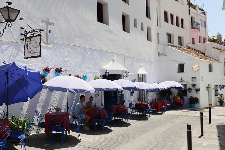 White washed building with blue plant pots on the wall and tables outside with blue umbrellas.