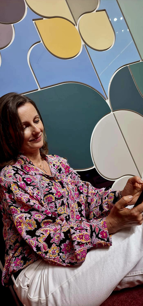 Glimlachende vrouw draagt bloemenblouse zittend voor kleurrijke abstracte muurkunst.