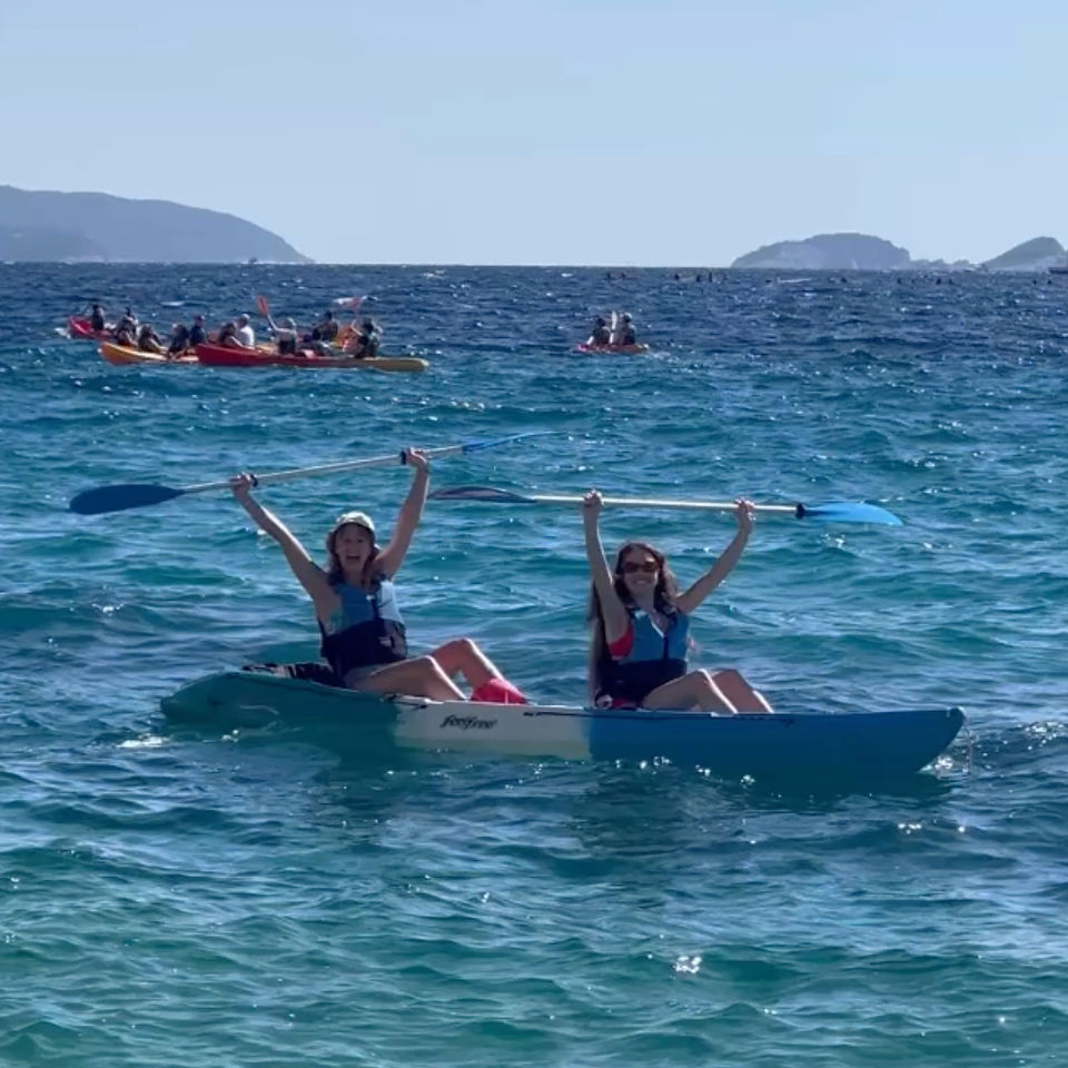 Two women sat in a blue kayak in the blue sea smiling and holding the paddles above their head