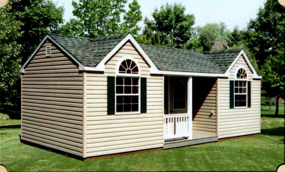 Quality Vinyl Cottage with double dormer design featuring tan siding, white trim, forest green shutters, palladian window with sunburst design, covered porch with white railing, architectural shingle roof, displayed on grass with natural backdrop