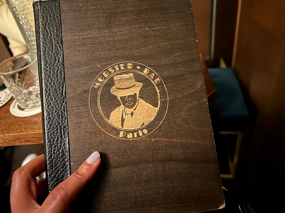 Hand holding a menu with a wooden cover featuring a "Mobster Bar Paris" logo. Background has a glass and coaster on a wooden table.