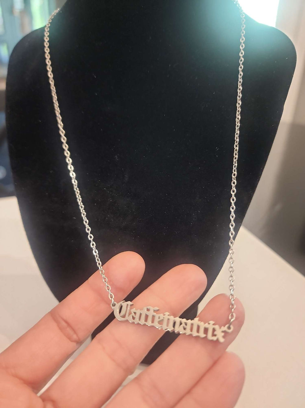 This is the Caffeinatrix stainless steel necklace that says the word Caffeinatrix in Old English. In this image someone is holding the lower portion of the necklace with their fingertips in order to show a size reference.