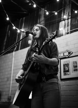 Allyn Raney performs live with her guitar in a black-and-white photo under string lights.